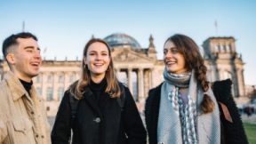Drei Studierende stehen lächelnd auf der Wiese vor dem Berliner Reichstag.